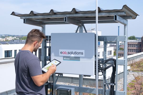 eco2solutions | Mann checkt mit Tablet die Photovoltaik-Anlage auf dem Dach und steht vor einer technischen Anlage mit eco2solutions-Aufkleber